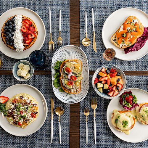 A table holds plates of various brunch foods, including waffles, eggs, avocado toast, pasta, fruit salad, and muffins, with drinks and cutlery.