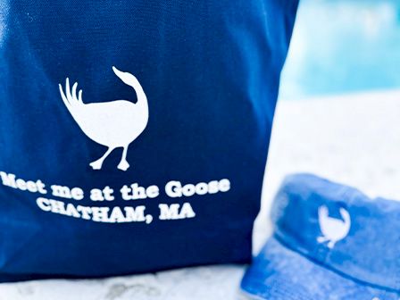 The image shows a navy blue bag and cap with a white goose logo and the text 