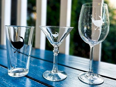 The image shows three types of glassware on a blue table: a pint glass, martini glass, and wine glass, each with a bird design on them.