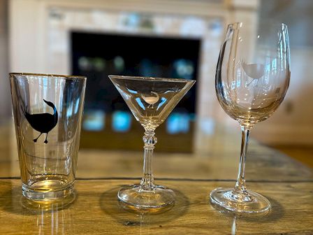 The image shows three different glasses on a wooden table, each with a bird design. The glasses include a tumbler, a martini glass, and a wine glass.