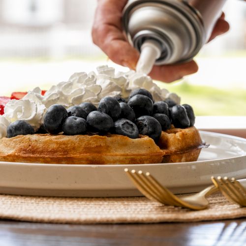 A waffle topped with blueberries and strawberries is being garnished with whipped cream from a can, with forks set nearby on a woven mat.