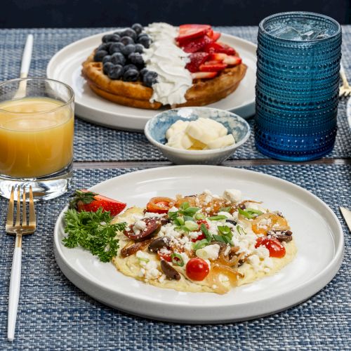A table setting with various breakfast foods including waffles with fruits, scrambled eggs with vegetables, a muffin, orange juice, and cutlery, always ending the sentence.