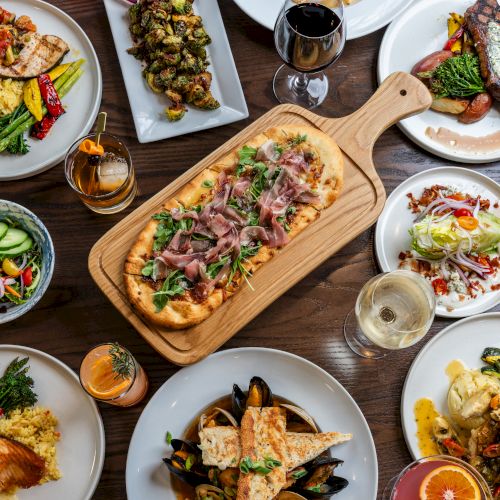 An assortment of dishes including pizza, steak, seafood, and salads on a wooden table, accompanied by various drinks like wine and beer.