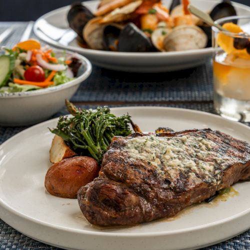A plate with steak, potatoes, and greens, a bowl of salad, seafood dish, and a cocktail on a blue mat.