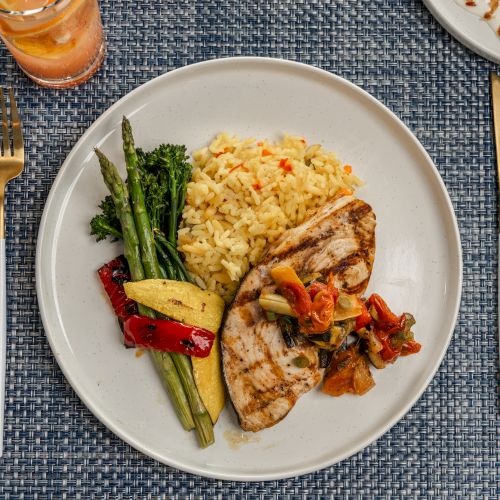 A plate with grilled chicken, vegetables, and yellow rice on a blue woven placemat, accompanied by a drink and utensils.