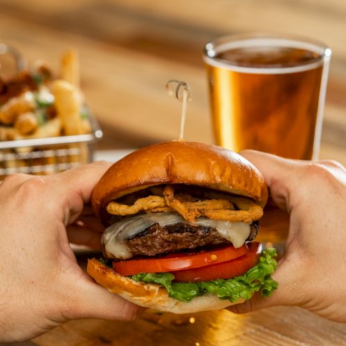 A person holds a loaded burger with lettuce, tomato, cheese, and crispy onions, with fries and a glass of beer in the background.
