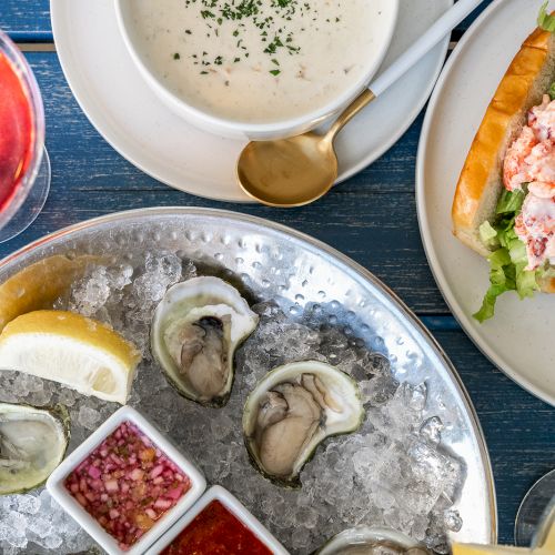An assortment of seafood dishes, including oysters on ice with sauces, lobster roll, a bowl of chowder, and a glass of rosé wine.