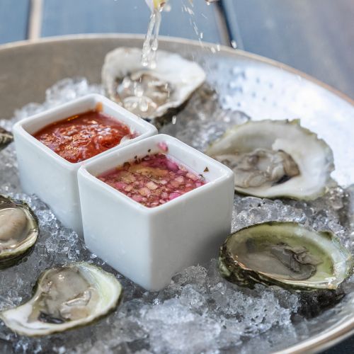 A plate of oysters on ice is being served with various sauces in square bowls, and lemon juice is being squeezed over the oysters.